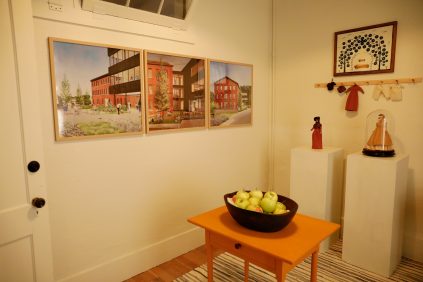 Room with framed architectural drawings on the wall, a table with a bowl of apples, and decorative items on pedestals.