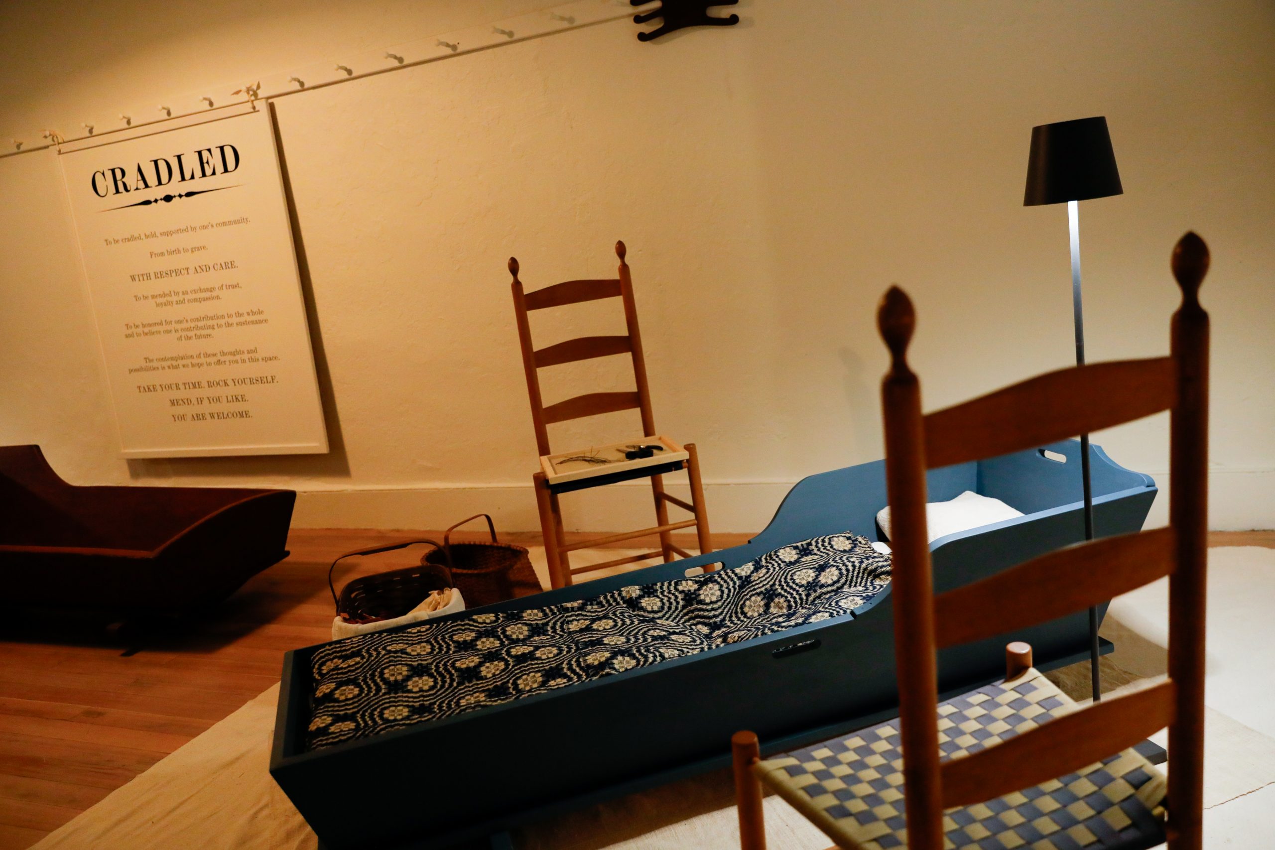 A museum exhibit featuring cradle displays, wooden chairs, and a patterned quilt. A wall sign titled 