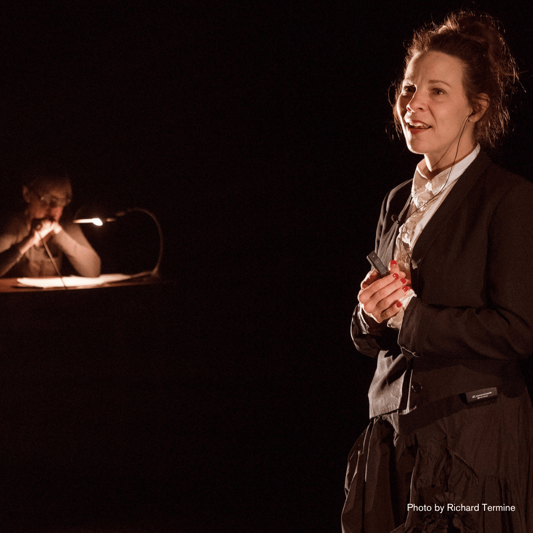 Suzanne Bocanegra and Lili Taylor on a dark stage. Lili wears her hair in a ponytail and is wearing a men's suit. Suzanne sits at a desk in the background, lit by a small light.