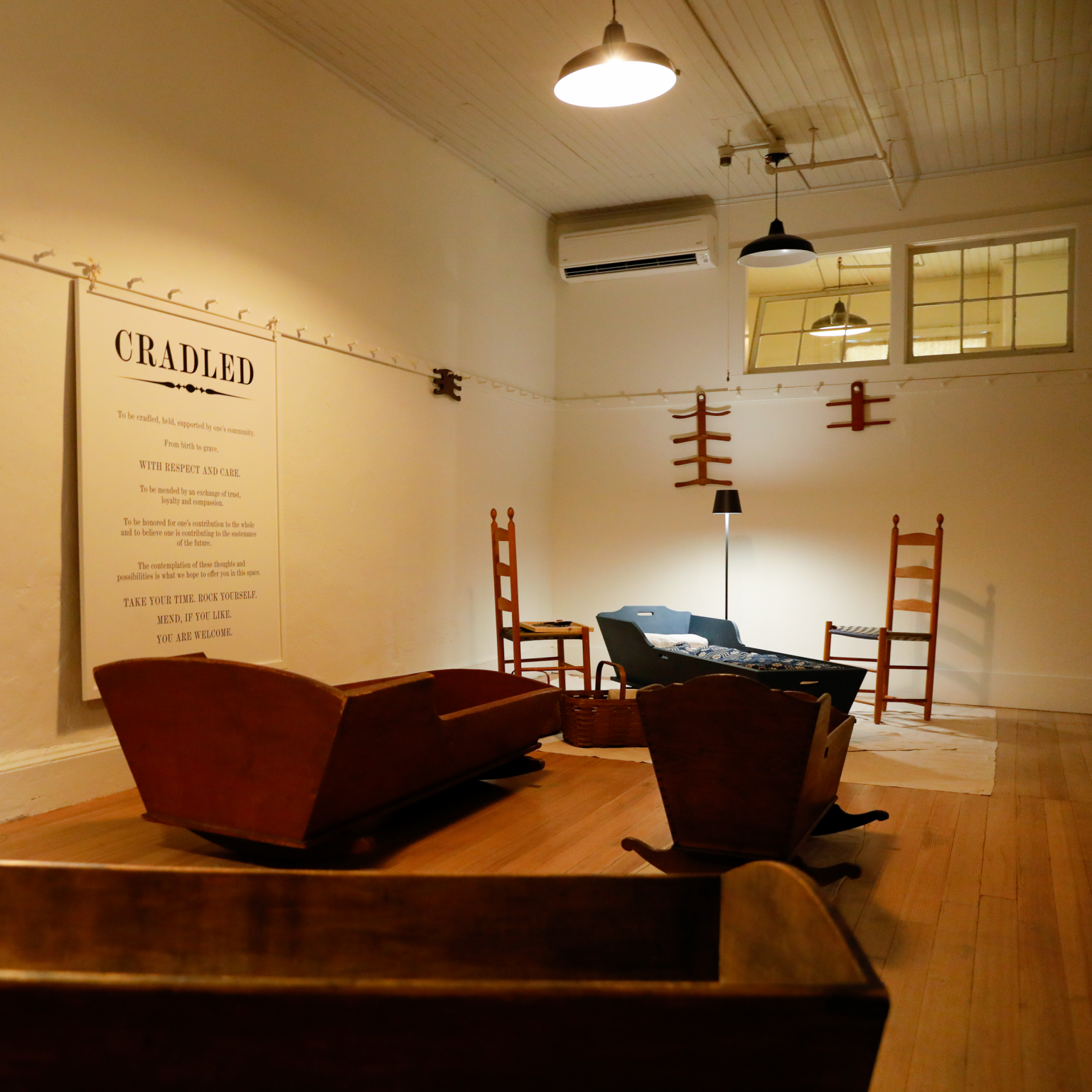 A photograph of the CRADLED exhibition featuring adult-size and child-size cradles, shaker chairs, and shaker hangers