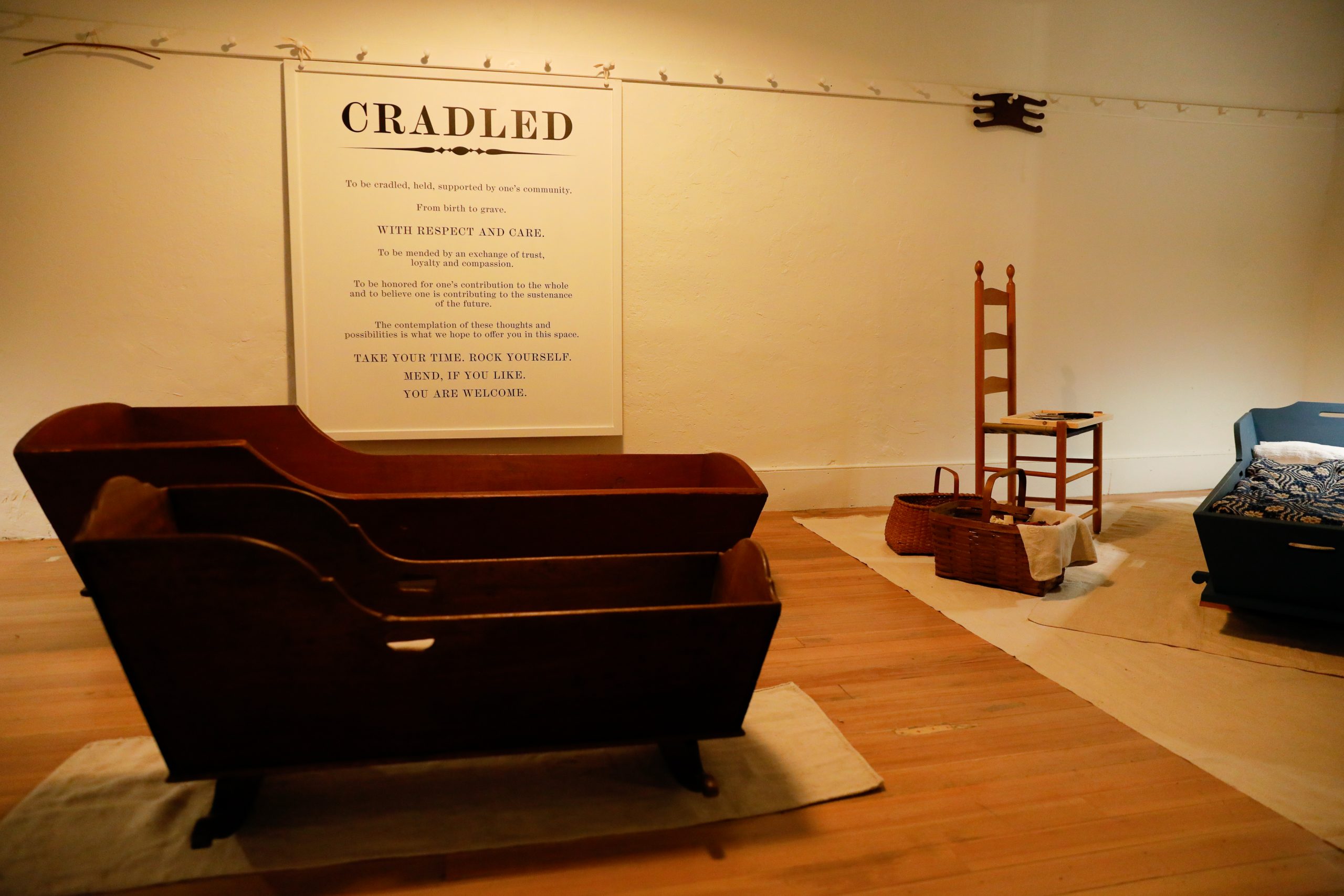 Exhibit with wooden cradles, a tall chair, and woven baskets. A large sign titled 