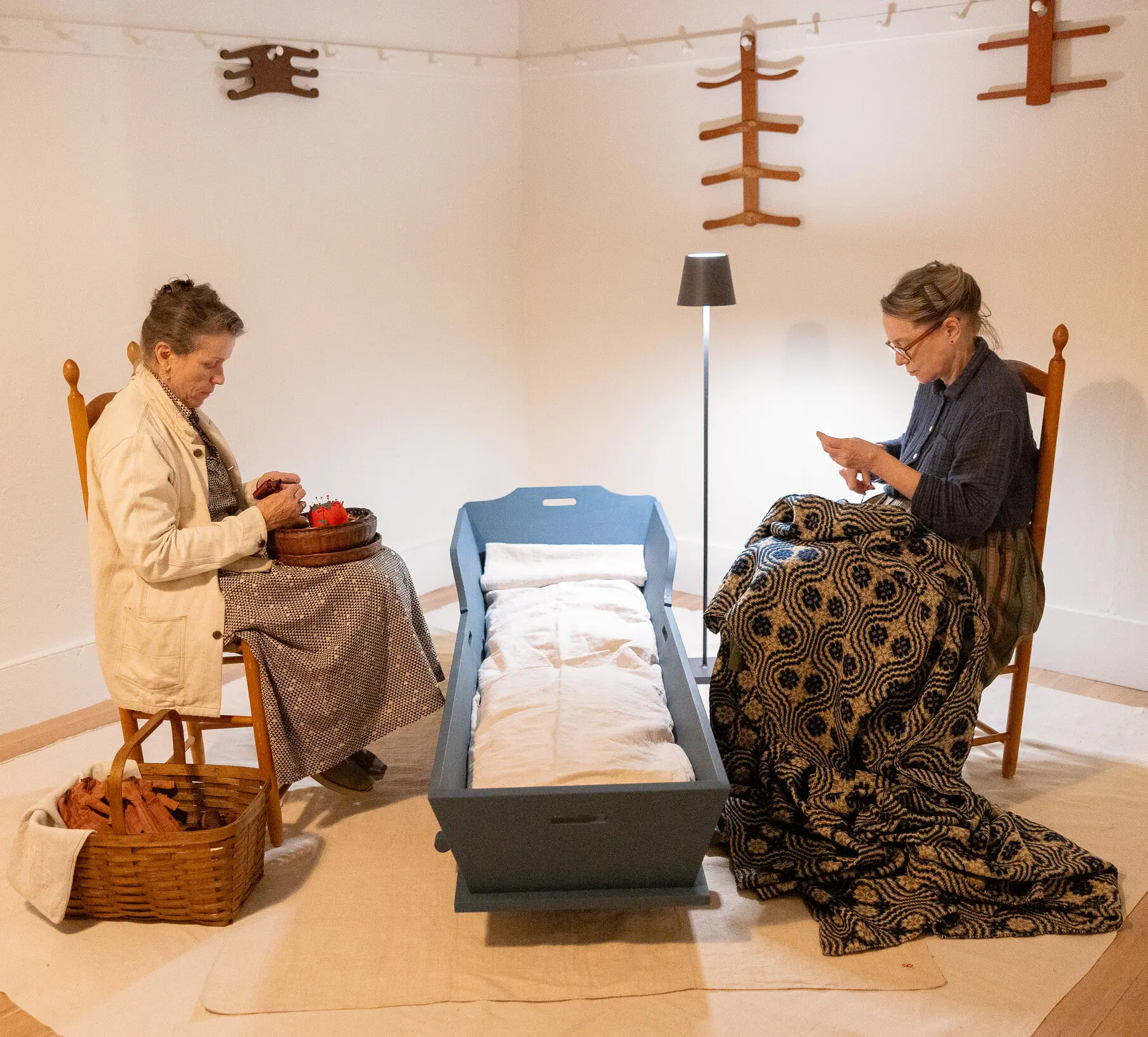 Two women sit on chairs facing each other in a room, engaged in sewing activities. Between them is a blue wooden cradle with a blanket. A lamp stands nearby. Wall decorations are visible in the background.