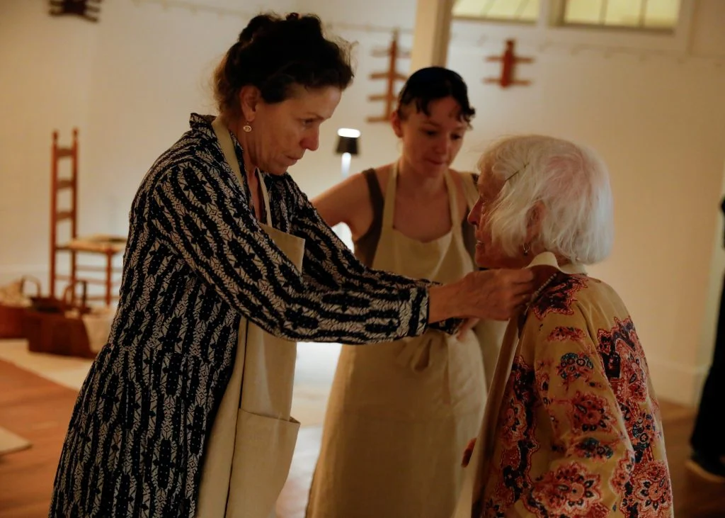 Three women are interacting in a room with wooden furniture. One is adjusting the jacket of an elderly woman in a patterned top, while another looks on with a slight smile.