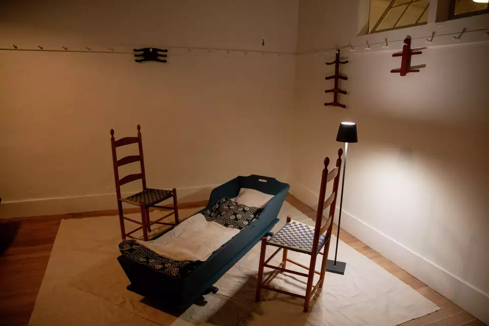 A dimly lit room displays a cradle, two chairs, a standing lamp, and red wooden decorations on the walls.