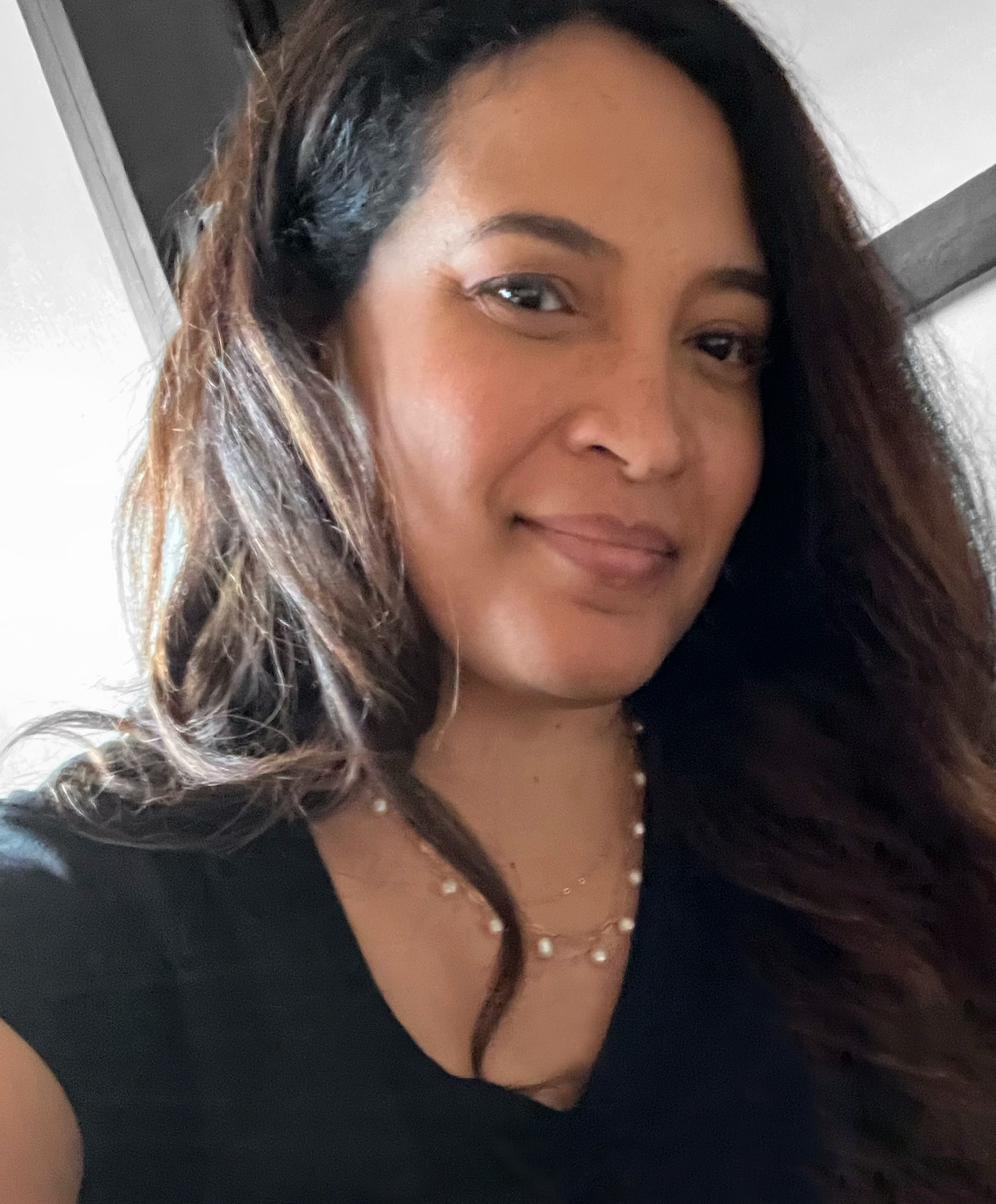 A woman with long brown hair and a black top smiles slightly at the camera. She is indoors, and a necklace with small pearls is visible.